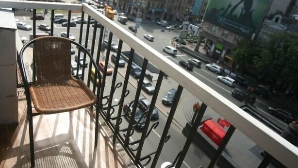 Un balcón con sillas de mimbre, vista de una calle concurrida con coches y edificios, y una caja roja en la barandilla.