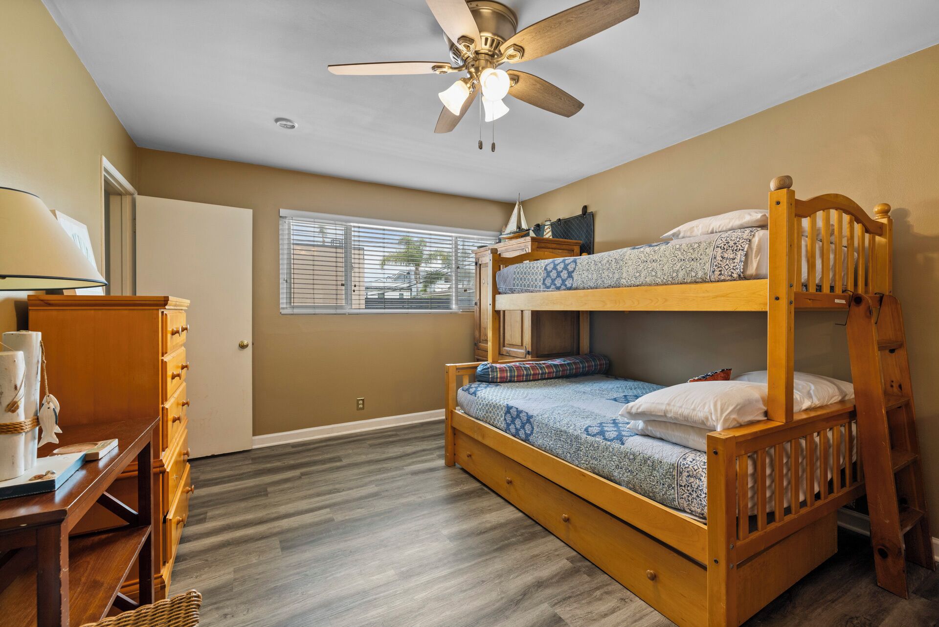 A bunk bed with a ceiling fan, a desk with a lamp, and a window with blinds.