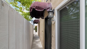 A narrow alleyway with a white wall on one side and a black door on the other. A purple awning hangs over the door, and a lantern is mounted above it. - Cozy Condo With New AC, Steps from Balboa Ferry and Peninsula Beach Access (Balboa Island, Newport Beach)