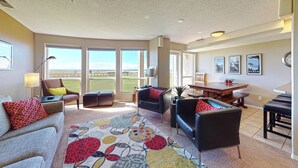 A spacious living room with a large window, a dining area, and a view of the outdoors. - OCEAN FRONT: DESTRESS RX: Seaside Vacation Homes, Inc Managed (Seaside)
