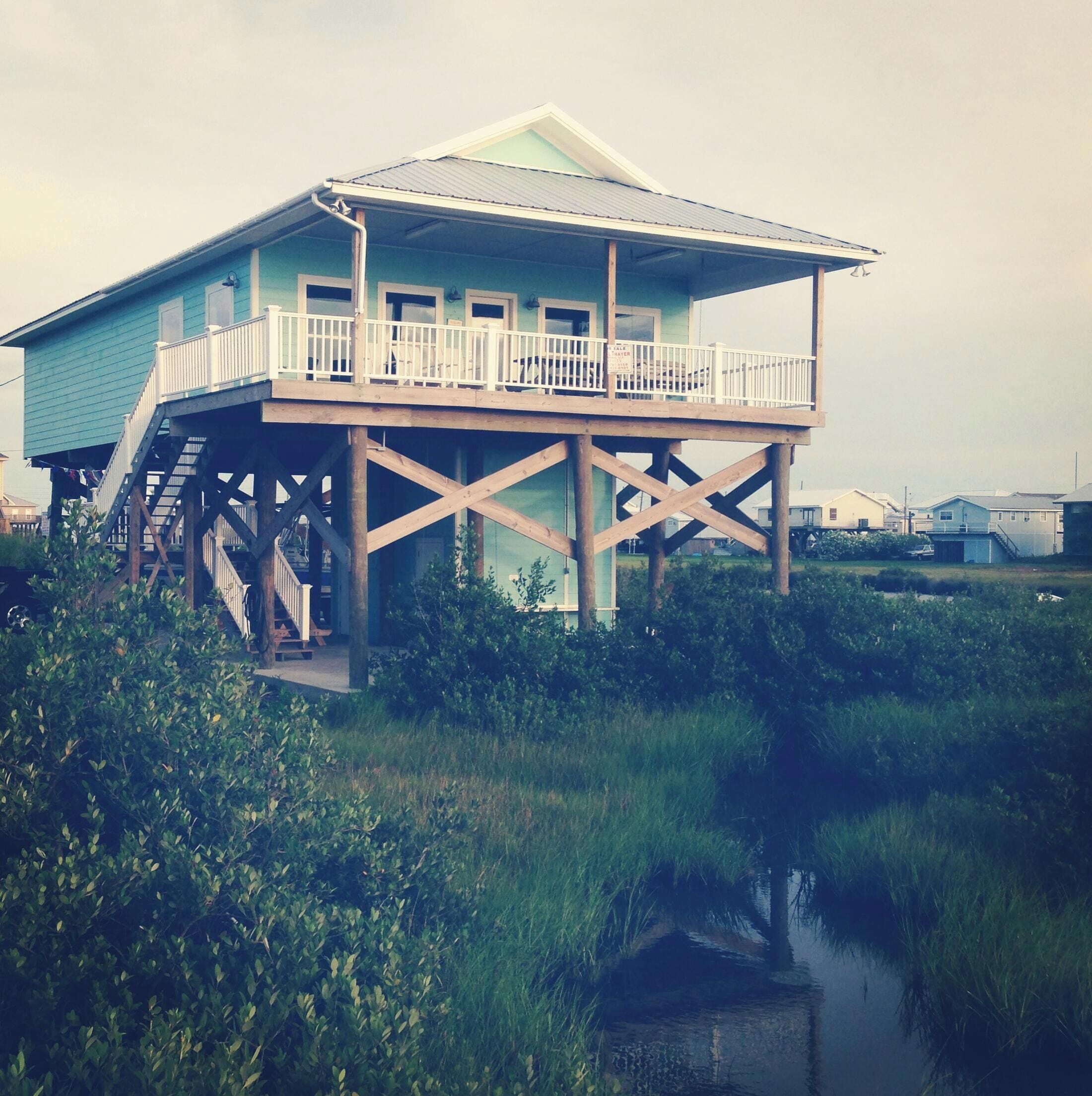 A house with a balcony on stilts over water, surrounded by greenery.