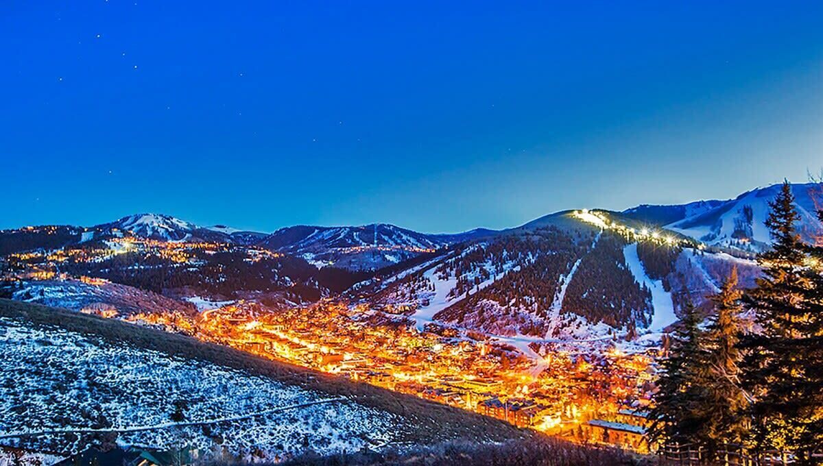 Paisaje montañoso nevado de noche con un pueblo iluminado por luces.