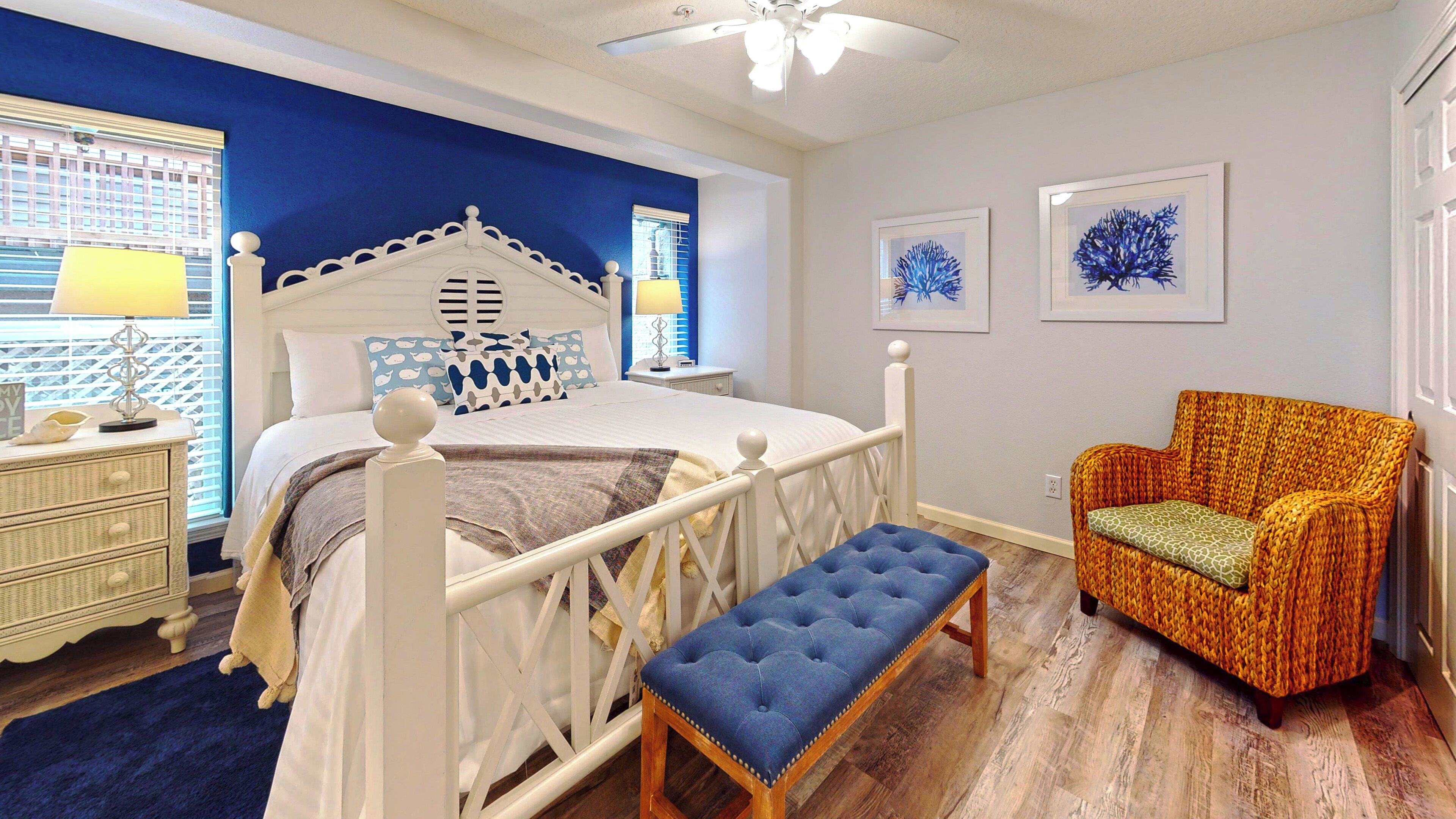 Quarto com cama branca, um banquinho azul, poltrona de vime e obras de arte na parede.