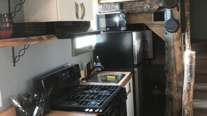 A compact kitchen with a stove, microwave, and refrigerator. There are wooden shelves with dishes and a rustic decor element. - Cozy "tiny home" Cabin with amazing views (Mountain View)