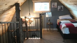 A cozy attic bedroom with a sloped ceiling, a bed with a red and white bedspread, a small table with a lamp, and a window with artwork. - Cozy "tiny home" Cabin with amazing views (Mountain View)