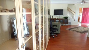 A spacious living room with a wooden floor, a glass cabinet with decorative items, a black office chair, a television, and a staircase with a red door. - A Foresthill Retreat (Foresthill)