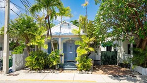 Une maison d’habitation dotée d’un porche à l’avant, entourée de plantes tropicales et d’une clôture blanche en bois.