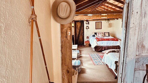 A rustic wooden interior with a narrow hallway leading to a bedroom. Visible features include a wooden ceiling, a hat on the wall, and a bed with a patterned bedspread.