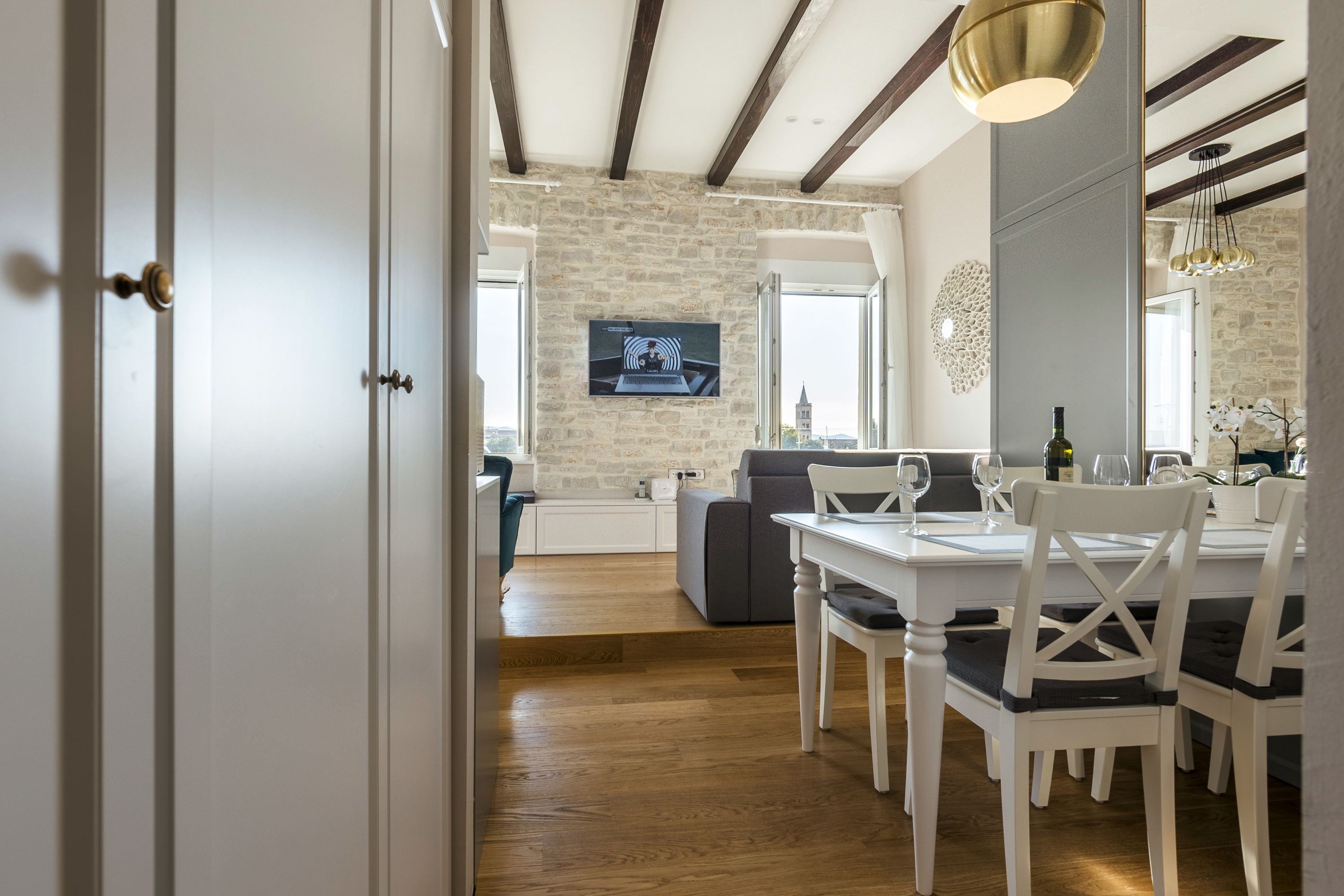 A dining area with a white table and chairs, a stone wall, and a television mounted on the wall.