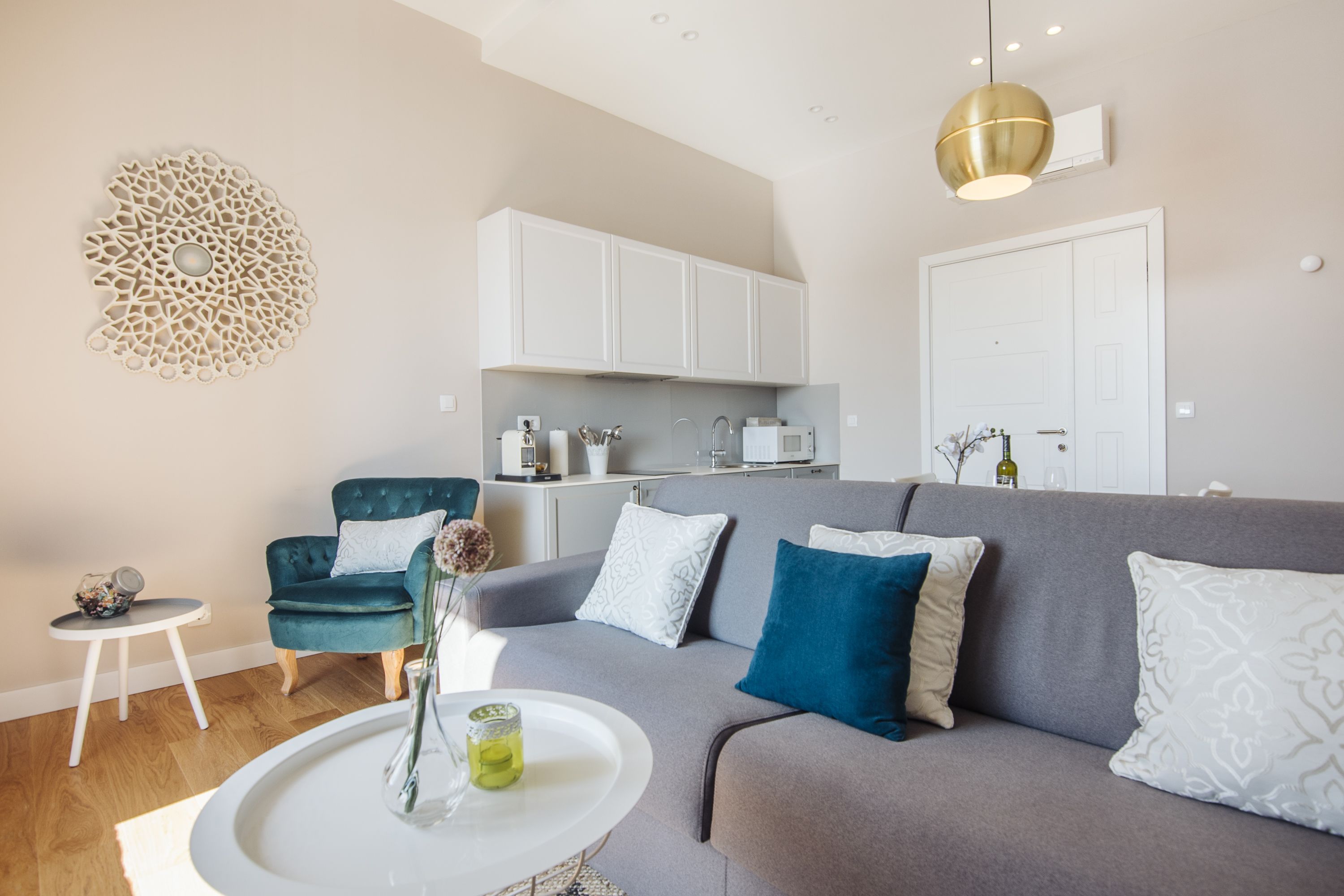 A modern living room with a grey sofa, a small white coffee table, and a teal armchair. There is a kitchen area with white cabinets and a microwave.