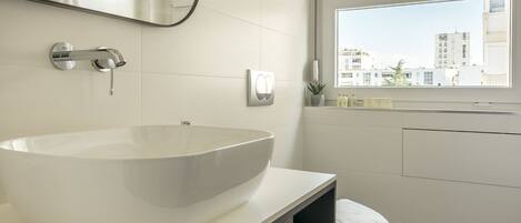 A modern bathroom with a large white sink, a mirror, and a window with blinds.