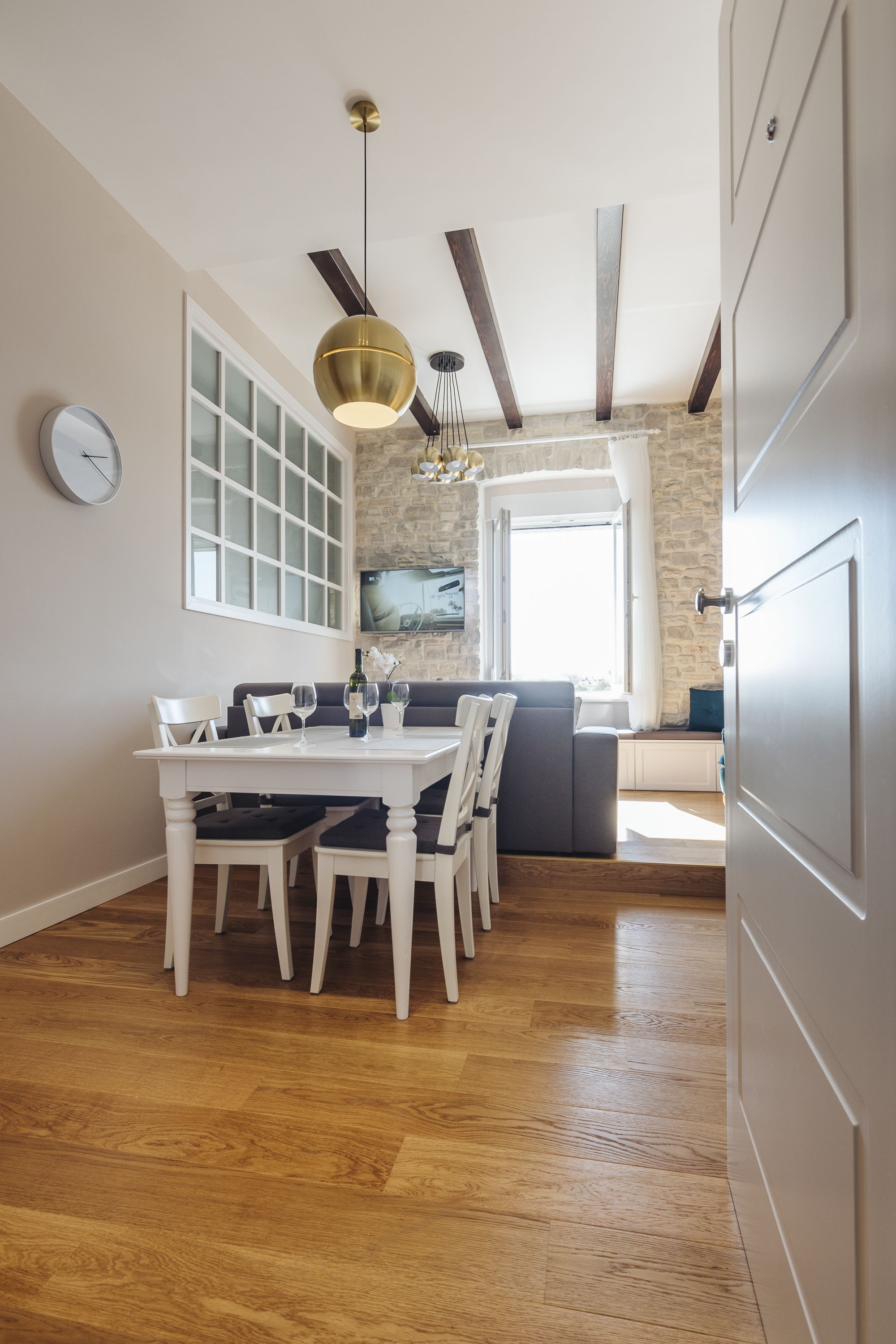 A dining area with a wooden table, white chairs, and a pendant light. There is a stone wall and a window with a view.