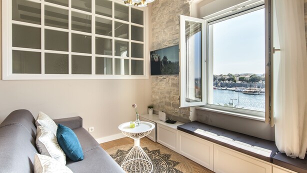 A cozy room with a gray sofa, a small white table with a vase, a large window with a view of a harbor, and a stone wall. - Magic View Apartments (Zadar)