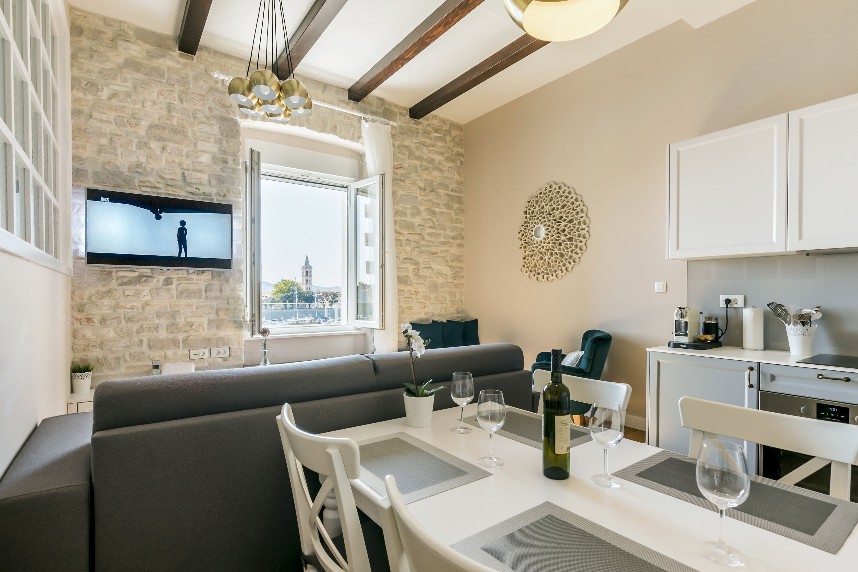 Una sala de estar moderna con pared de piedra, un televisor de pantalla plana, un área de comedor con mesa para dos, y una cocina con armarios y encimera.