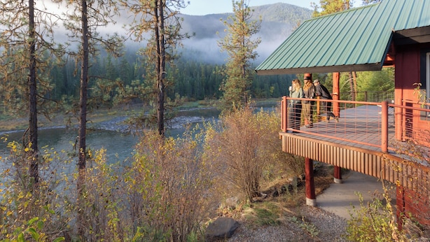 A wooden deck with a metal railing overlooking a river and forested mountains.
