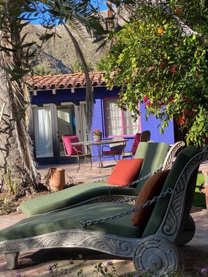 Un patio con una silla colgante, una mesa y sillas. Se ve un edificio con una pared azul y un techo de tejas al fondo.