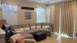 Ein Wohnzimmer mit einer Eckcouch, einem kleinen Tisch mit Lampe, einem Deckenventilator und einem großen Fenster mit Vorhängen.