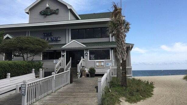 A two-story building with a sign reading "The Oceanic." It features a wooden deck leading to the entrance, a palm tree, and a view of the beach.