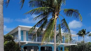 A two-story beachfront property with a pool, palm trees, and a red object on the sand.