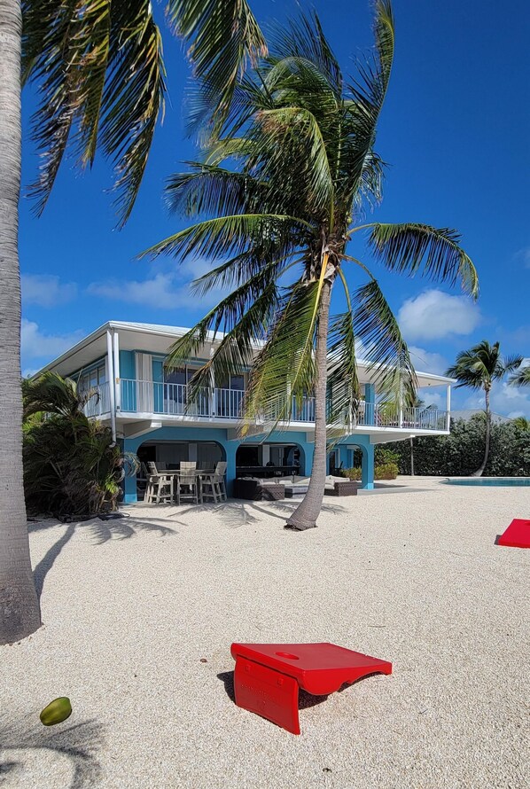 A two-story beachfront property with a pool, palm trees, and a red object on the sand.