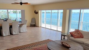 A spacious living room with a large window overlooking a body of water, a ceiling fan, and a dining area with chairs and a table.