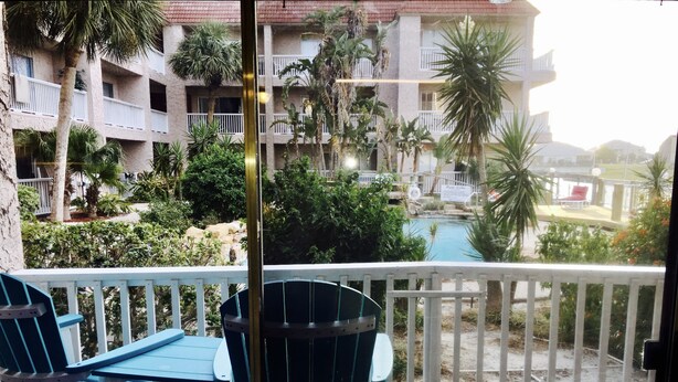 Un balcon avec deux fauteuils Adirondack bleus, une terrasse en bois et une vue sur une piscine entourée de palmiers et sur un immeuble doté de balcons.