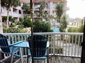 A balcony with two blue Adirondack chairs, a wooden deck, and a view of a pool surrounded by palm trees and a building with balconies. - Waterfront on Canal, Boat Slip, Close to Beach, Restaurants, Shopping & Fishing! (Corpus Christi)