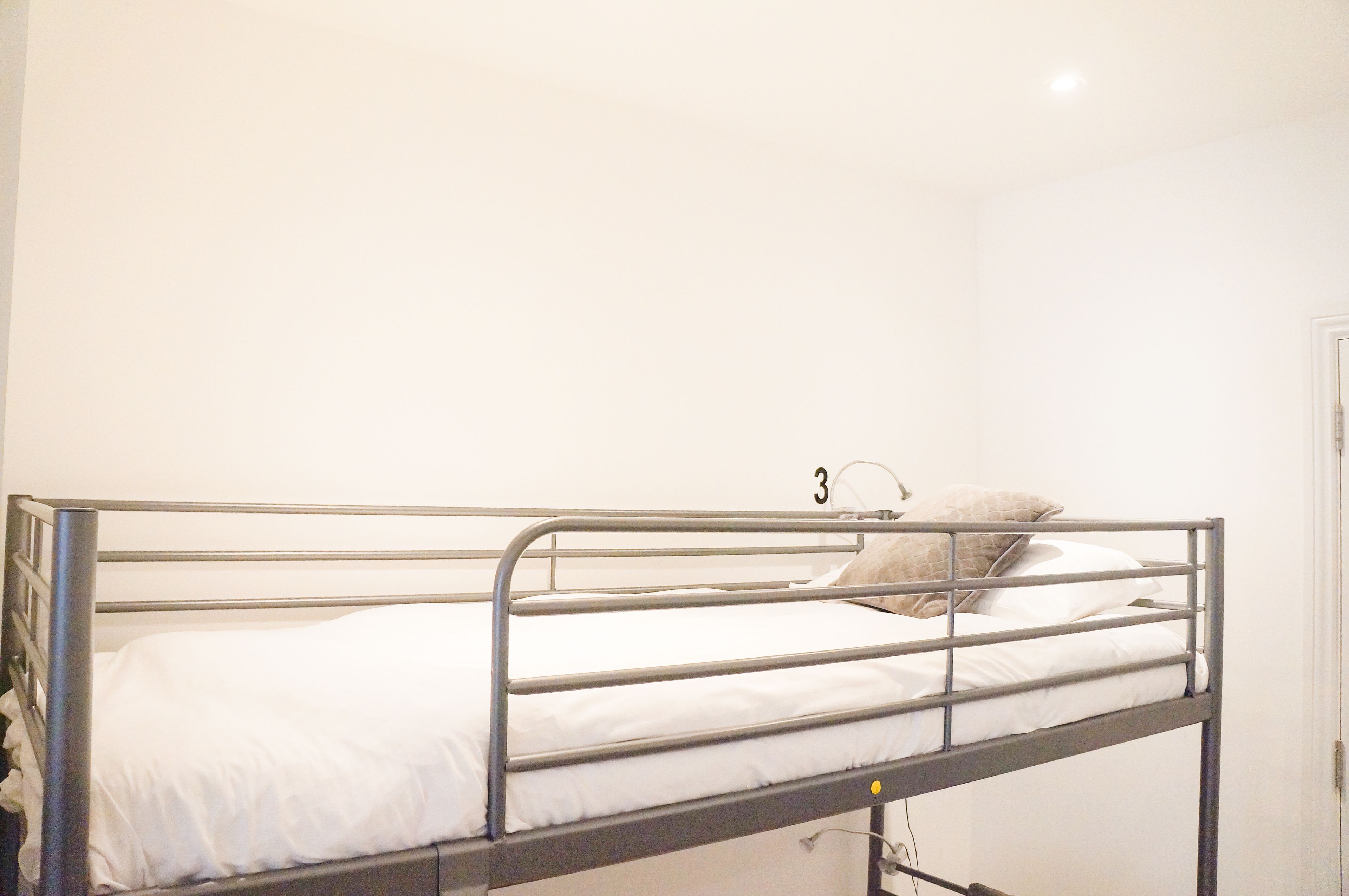 A single metal bunk bed with white bedding, a small bedside table, and a wall-mounted number plate.