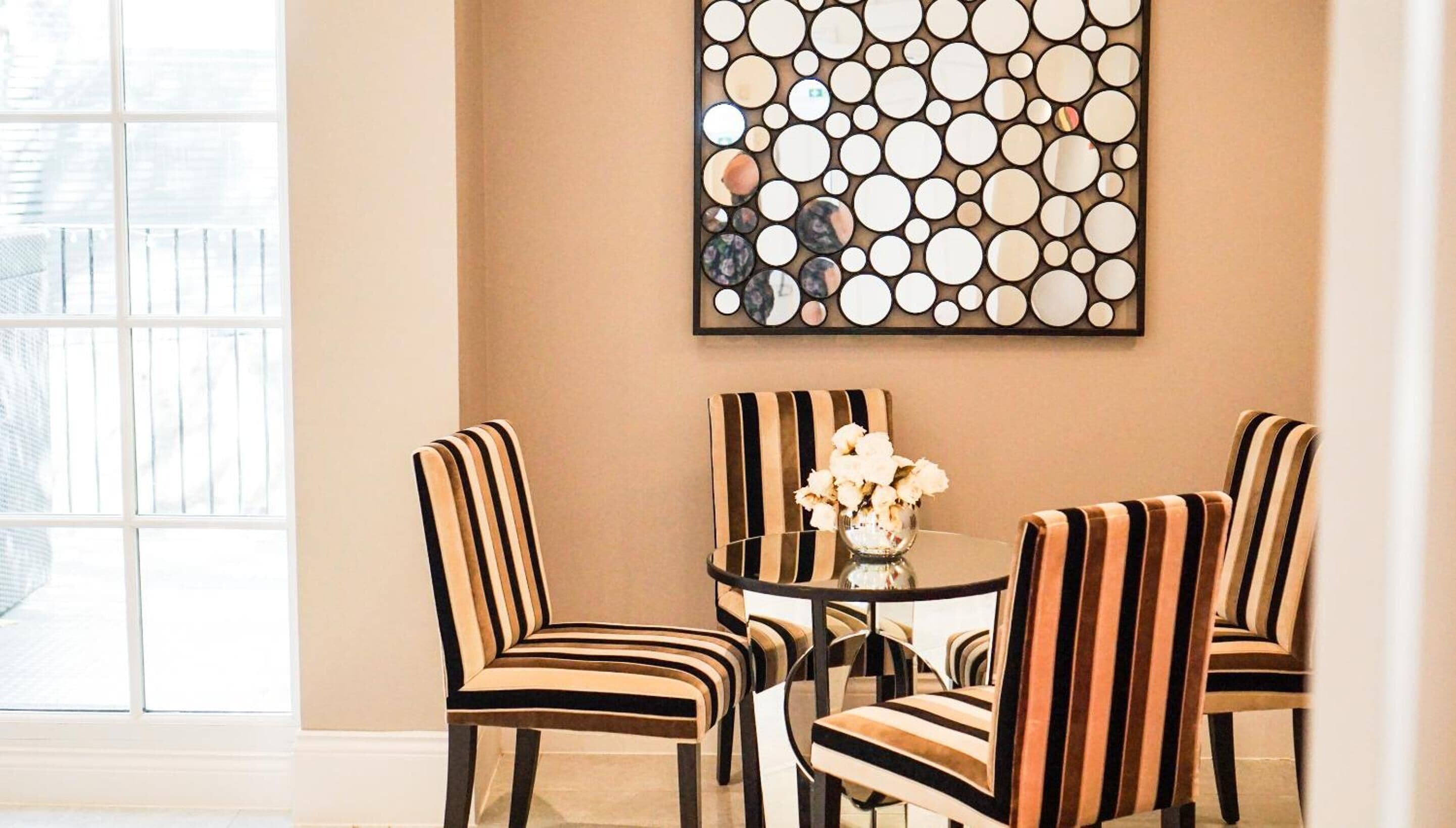 A dining area with striped chairs and a glass table, featuring a circular mirror artwork on the wall.