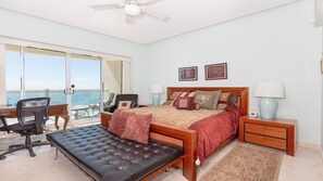 A bedroom with a bed, a desk, a chair, a bench, a nightstand, and a view of the sea.