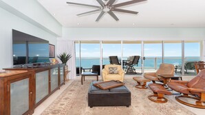 A spacious living area with a large window overlooking the ocean, a ceiling fan, and a wooden coffee table.