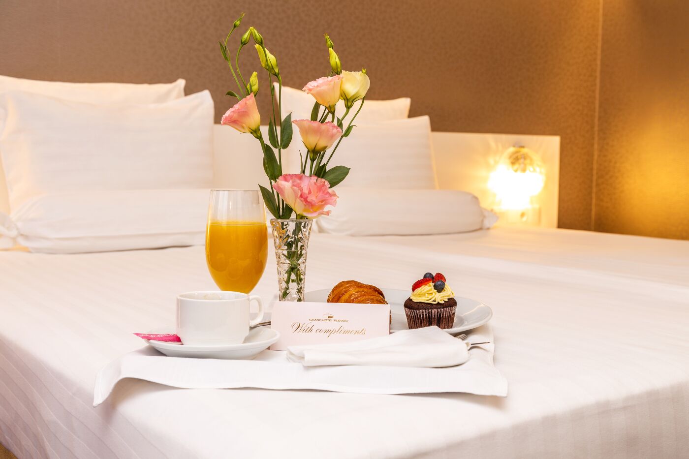 A neatly made bed with white linens, a glass of orange juice, a cup, a plate with a croissant and a cupcake, and a card that reads "Grand Hotel."