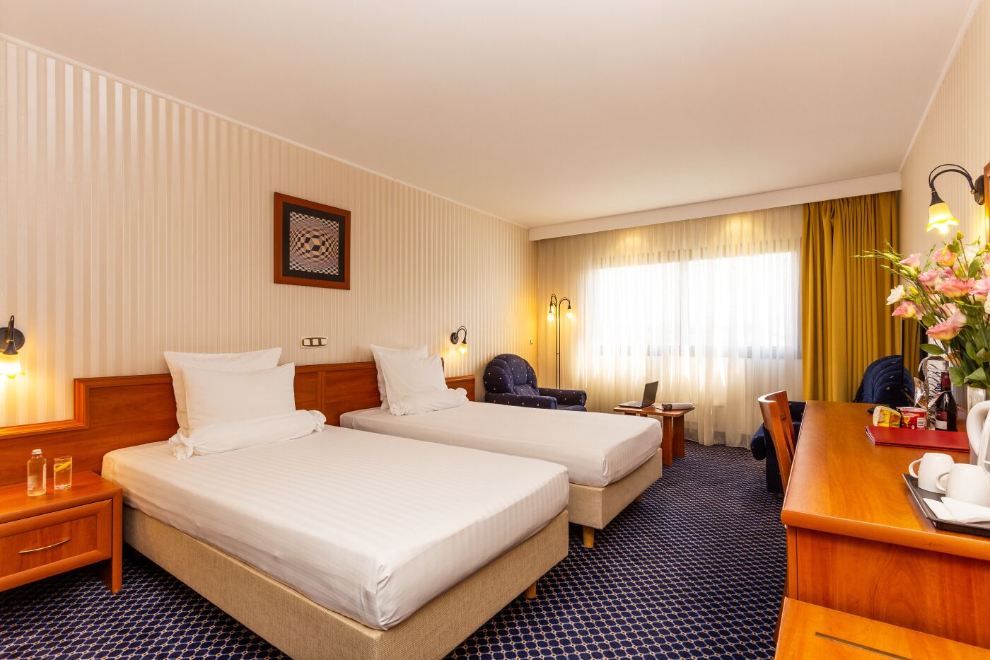 A hotel room with two beds, a desk, a chair, a laptop, a vase with flowers, and a window with curtains.