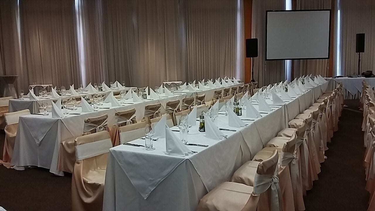 Un espai per a banquet amb llargues taules preparades per a un esdeveniment formal, amb estovatges blancs i fundes de cadira a conjunt.