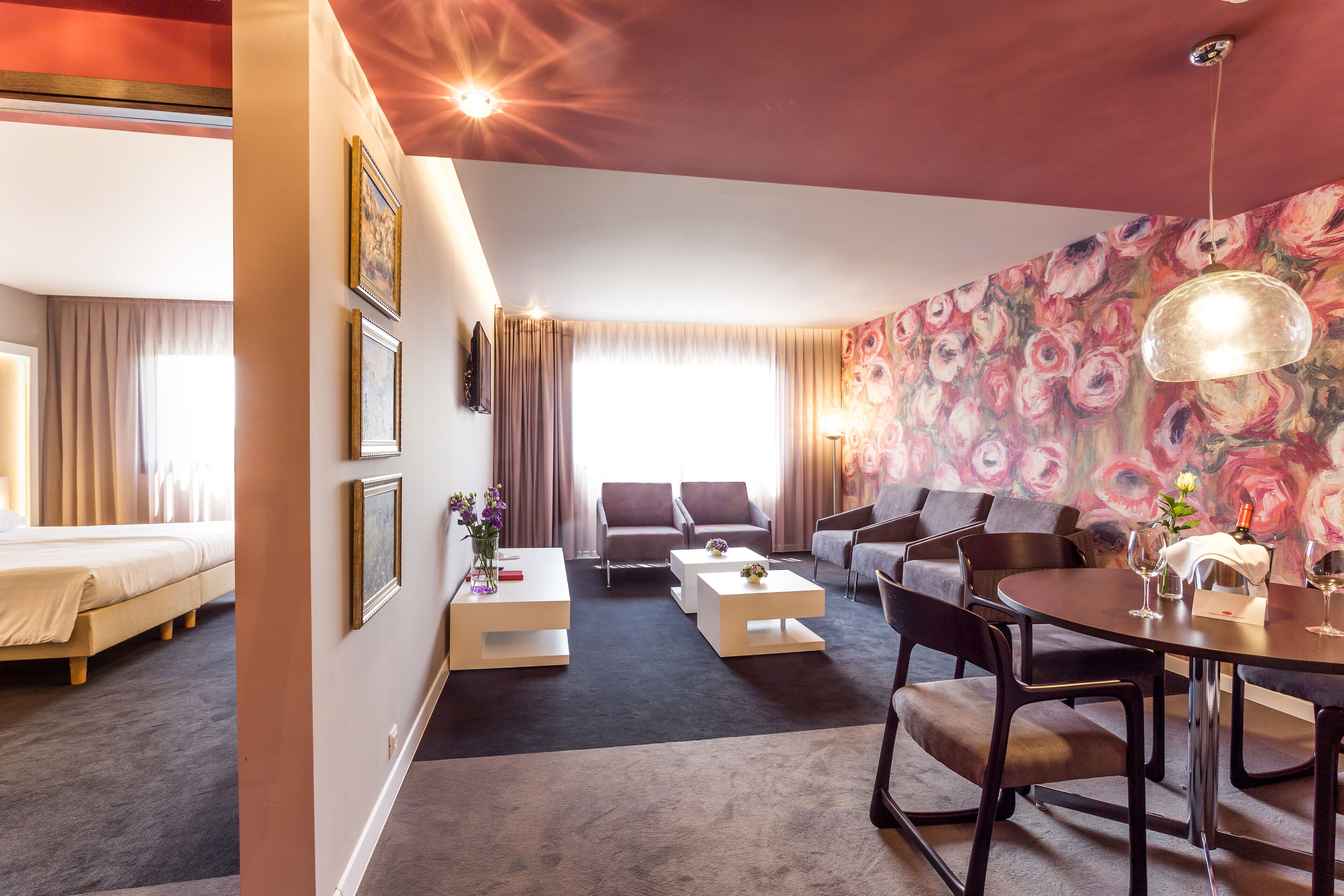 A modern hotel room with a bed, a sofa, a dining table, and a large floral mural on the wall.