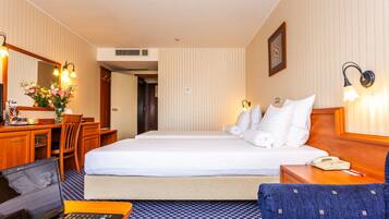 A hotel room with a bed, a desk with a laptop, a chair, a telephone, and a bedside table with a lamp.