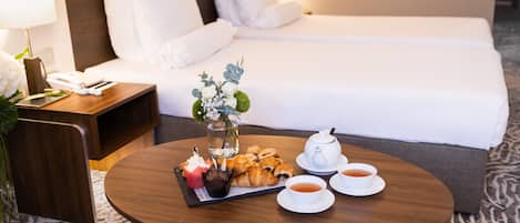 Camera d'albergo con un letto rifatto, un comodino con una lampada, un tavolino con pasticcini, un vaso con fiori e un servizio da tè.