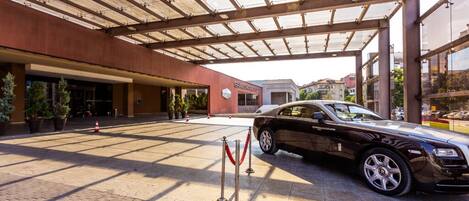 A luxury car parked under a covered entrance with a red carpet and stanchions.
