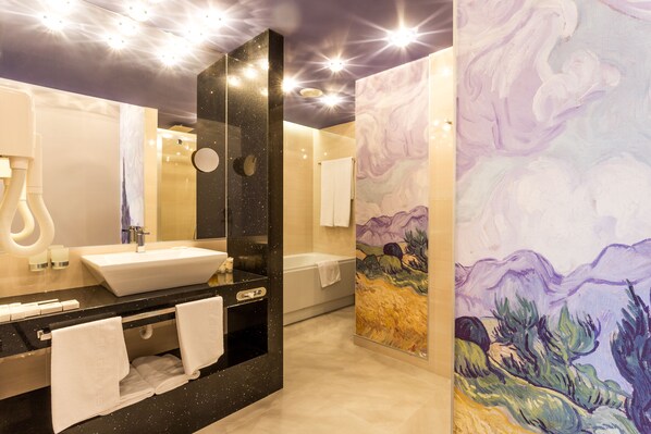 Un baño moderno con encimera negra, lavabo blanco y un espejo grande. Hay un cuadro en la pared y una bañera al fondo.