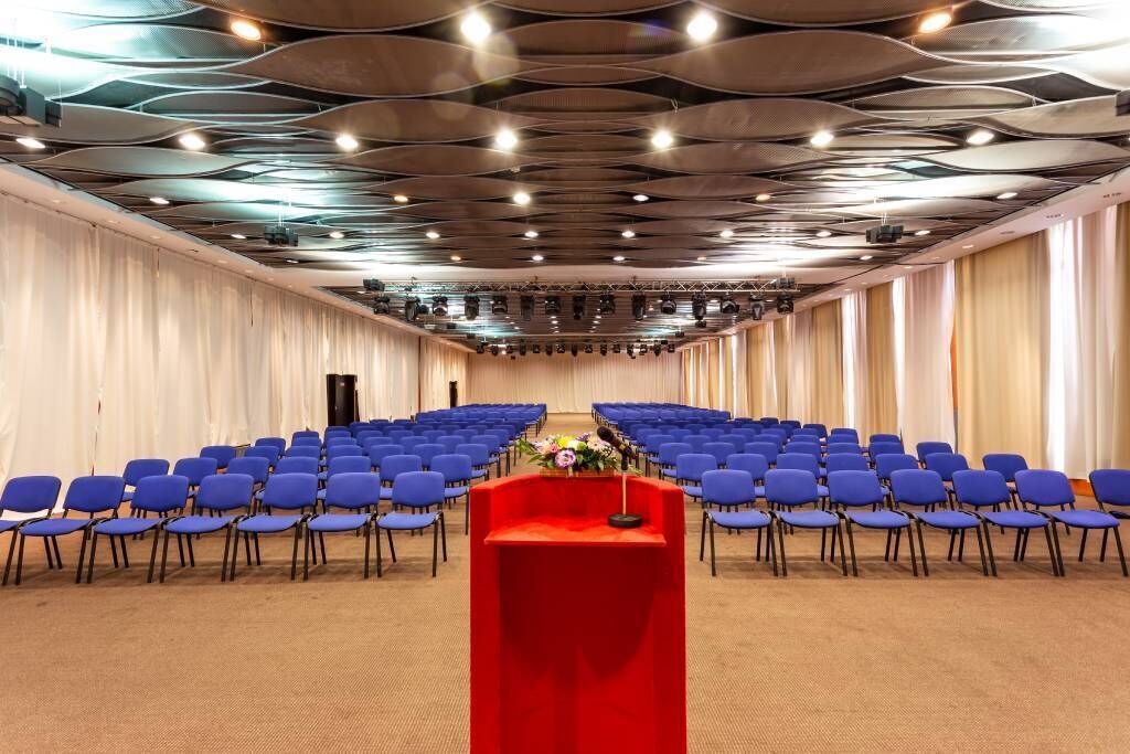 Un salón de conferencias con filas de sillas azules, un atril rojo y un diseño de techo moderno.