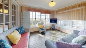 A cozy room with a sofa, a wicker chair, a TV on a stand, and large windows with curtains. - OCEAN VIEW: SAND HILL MANOR:  Seaside Vacation Homes, Inc. Managed (Gearhart)