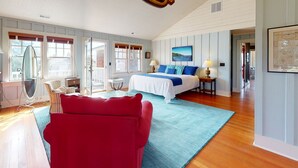 A spacious bedroom with a large bed, a red chair, and a view of the outdoors through the windows. - OCEAN VIEW: SAND HILL MANOR:  Seaside Vacation Homes, Inc. Managed (Gearhart)