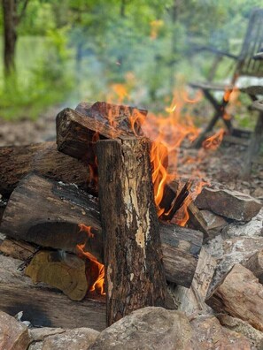 A campfire with logs and rocks, surrounded by trees and a chair in the background.
