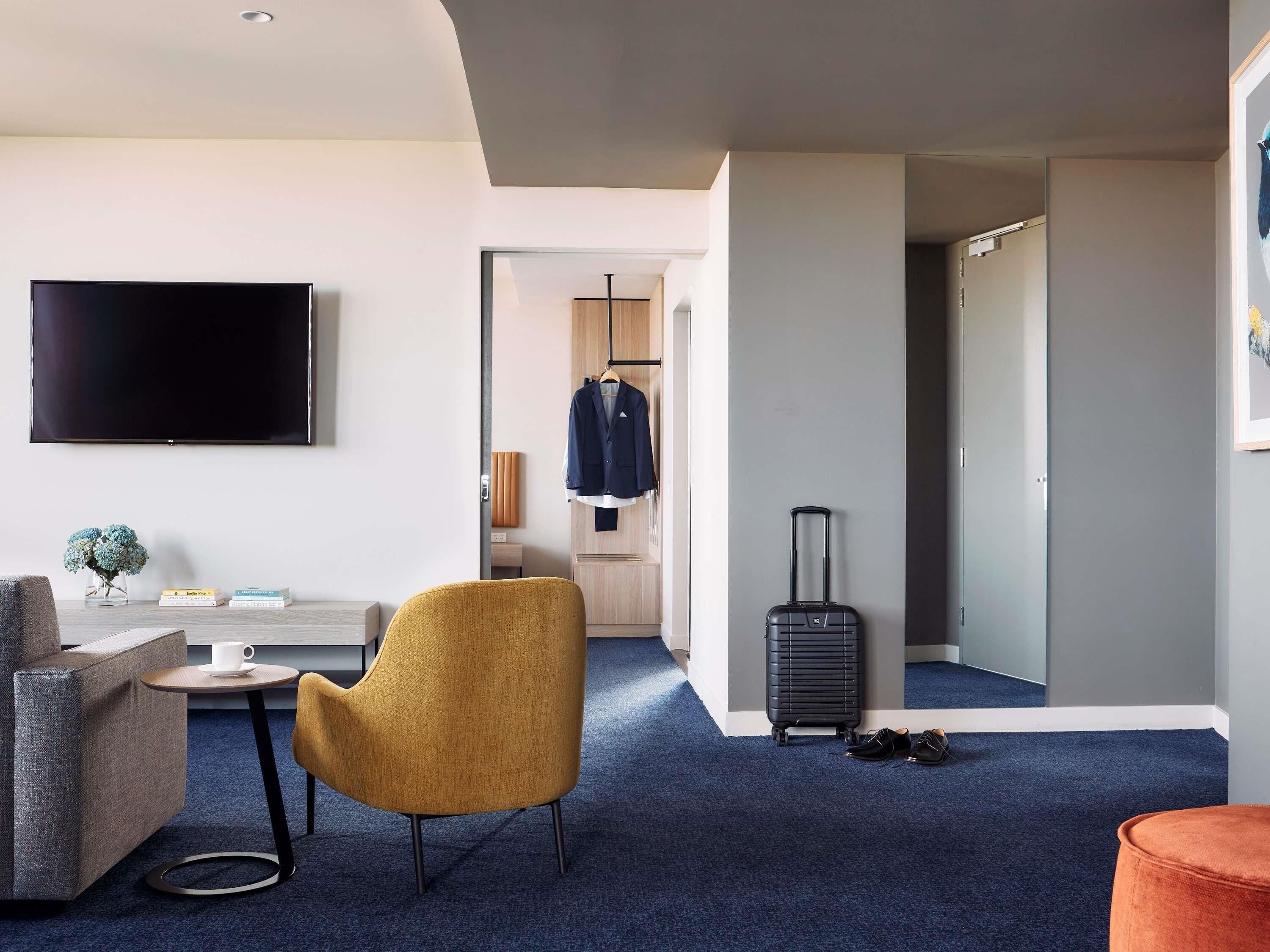 A modern hotel room with a flat-screen TV, a sofa, a small table, a chair, a suitcase, and a coat rack.