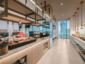 Un espace de restauration moderne dans un hôtel, avec un long comptoir, une variété de plats proposés et une vue sur l’extérieur.
