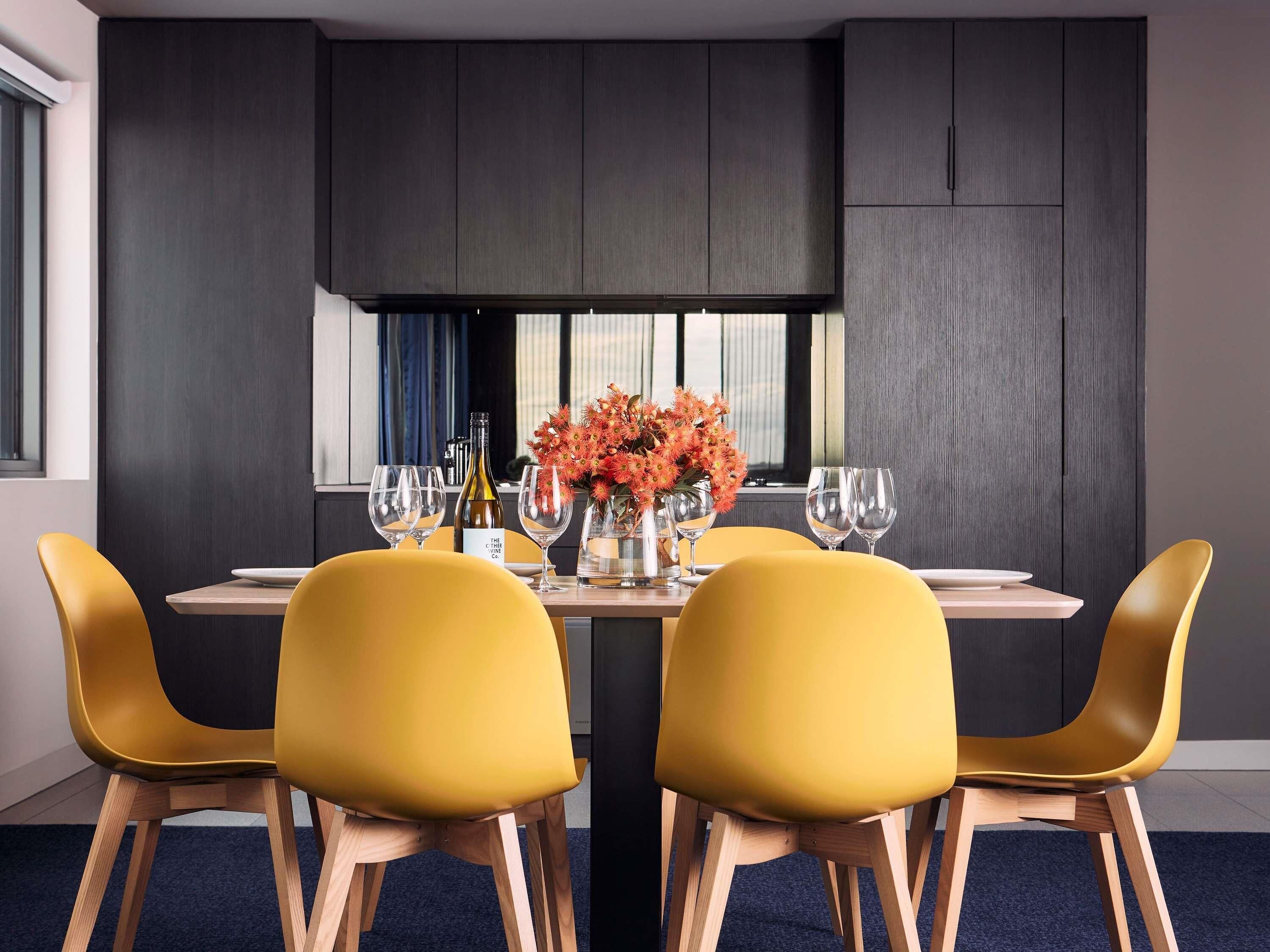 Un comedor moderno con una mesa dispuesta para dos personas, sillas amarillas, un jarrón de flores y copas de vino.