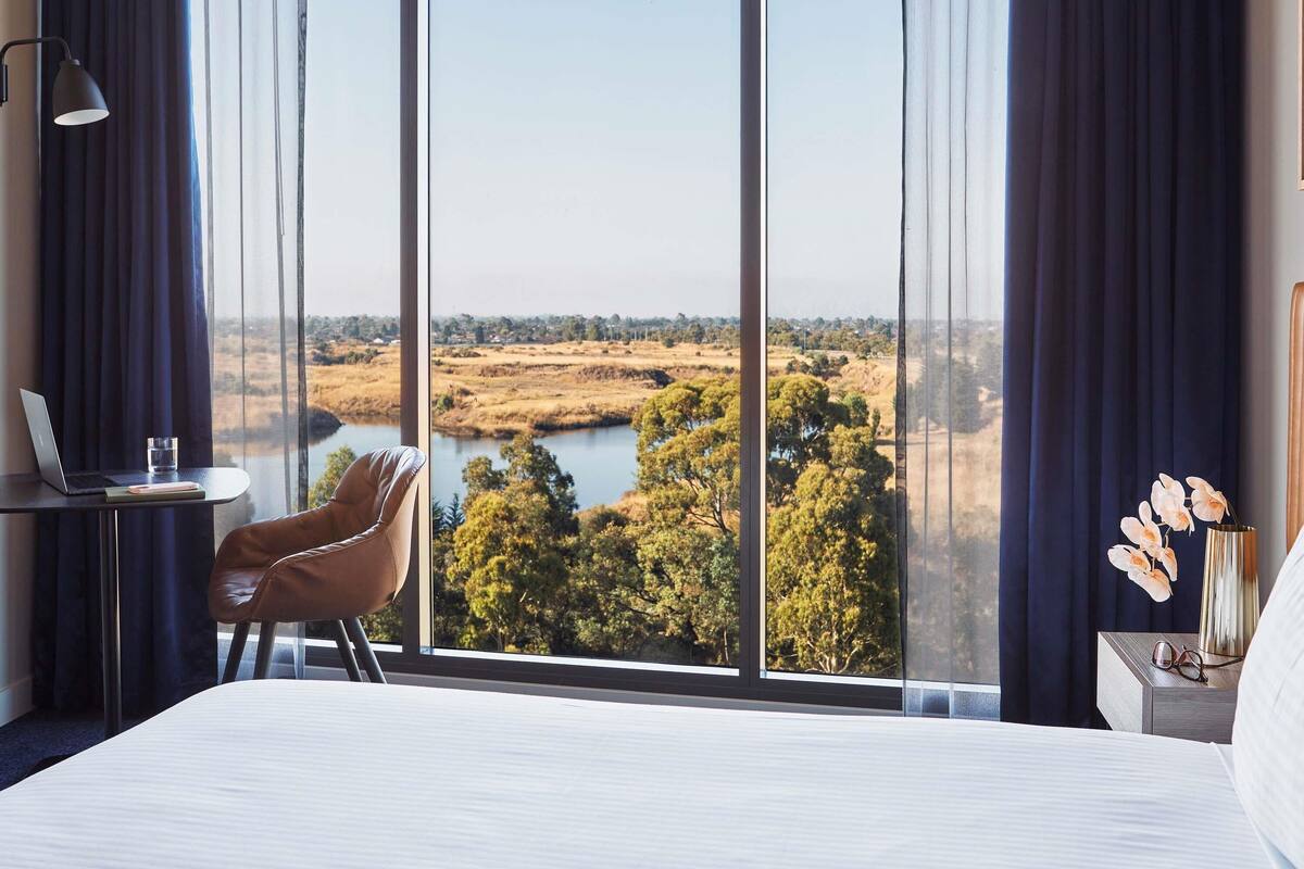 A hotel room with a large window, a bed, a desk with a laptop, a chair, and a nightstand with a vase of flowers.