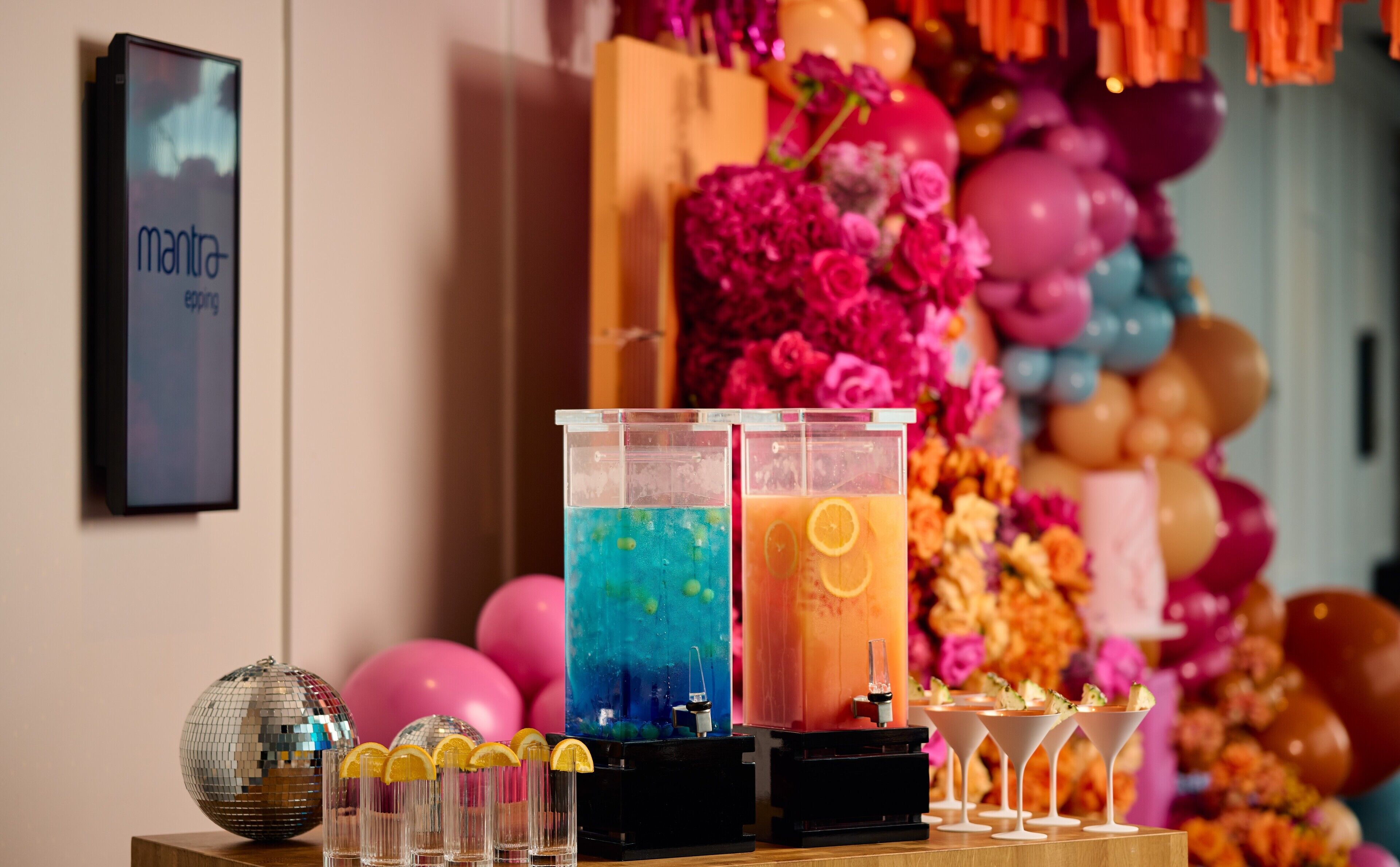 Zona de bar con dos dispensadores de bebidas, una bola de discoteca y una pared decorada con globos y flores.