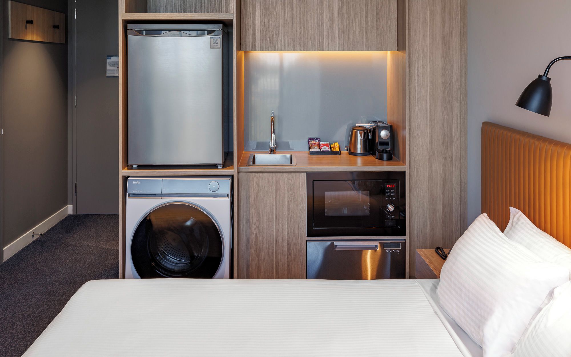 Habitación de hotel compacta con cama, lavadora, refrigerador, microondas y cafetera.
