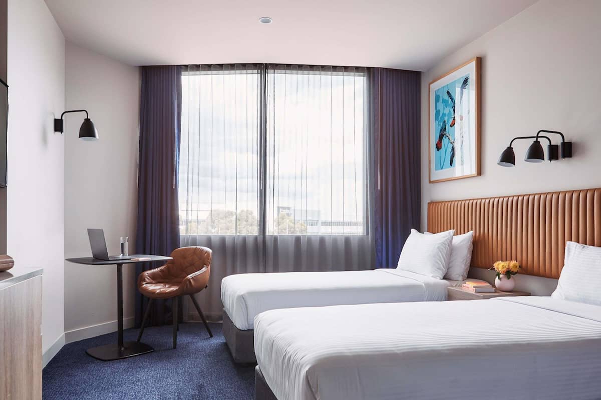A hotel room with two beds, a desk with a laptop, a chair, a window with curtains, and a painting on the wall.